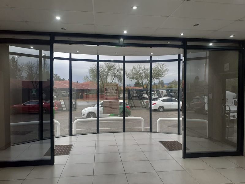 A bright commercial entrance lobby featuring large glass sliding doors, tiled flooring, and recessed downlighting, providing a clear view of the exterior parking area.