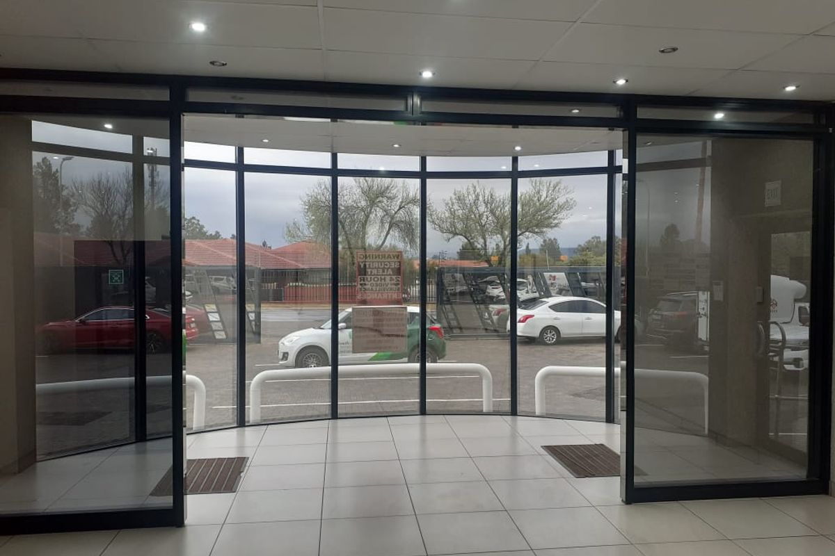 A bright commercial entrance lobby featuring large glass sliding doors, tiled flooring, and recessed downlighting, providing a clear view of the exterior parking area.