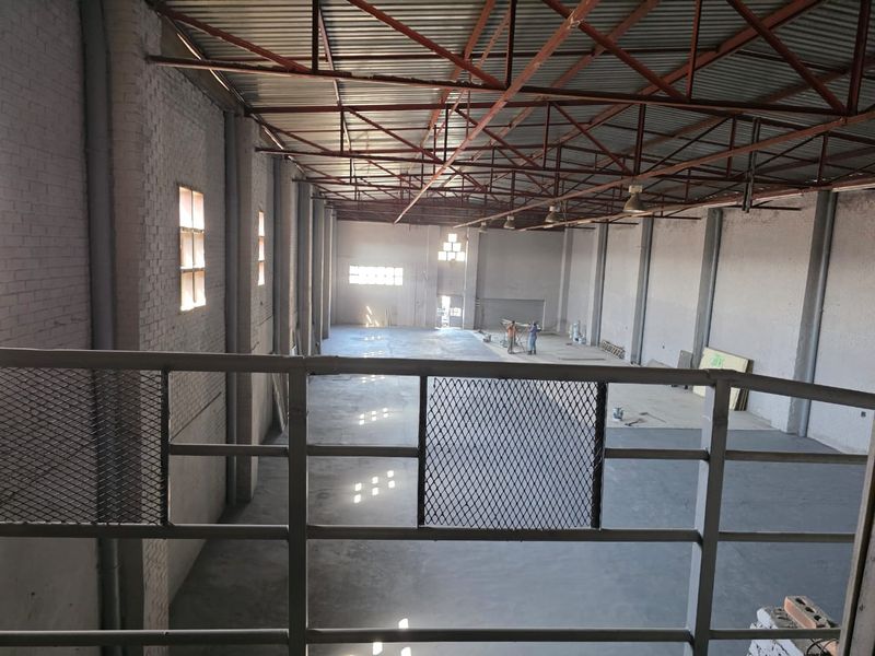 Spacious industrial warehouse area featuring high ceilings with exposed steel trusses, concrete flooring, and painted brick walls, viewed from a mezzanine level.
