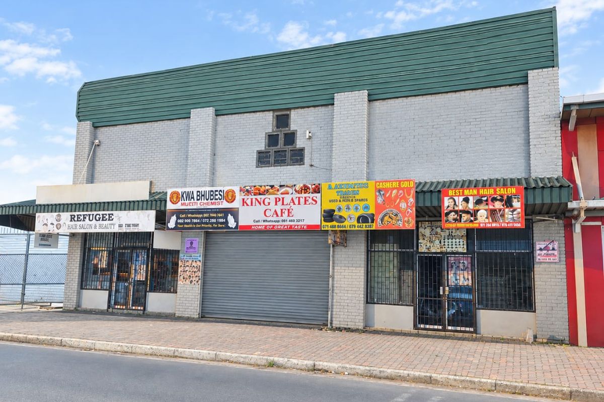 Front elevation of a multi-tenant industrial or commercial building featuring multiple storefronts, security shutters, and signage along a paved street.