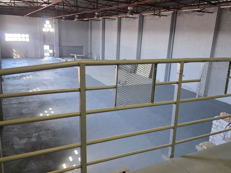 Spacious industrial warehouse interior featuring high ceilings with exposed steel trusses, concrete flooring, and a mezzanine level with metal safety railing.