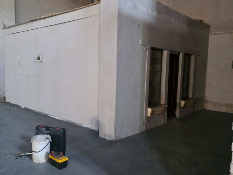 An industrial utility space featuring a standalone internal office structure, concrete flooring, and exposed brick walls, currently undergoing renovation.