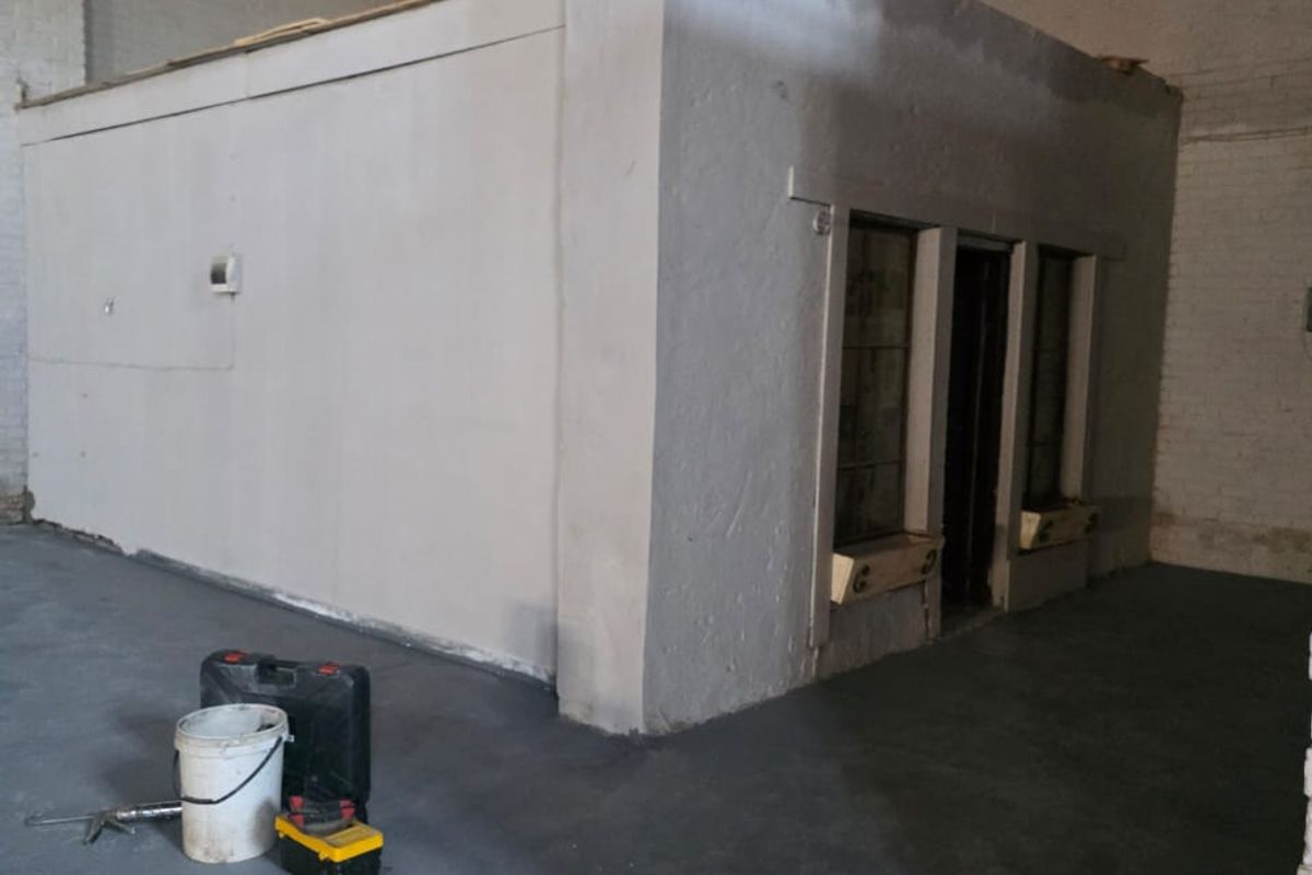 An industrial utility space featuring a standalone internal office structure, concrete flooring, and exposed brick walls, currently undergoing renovation.