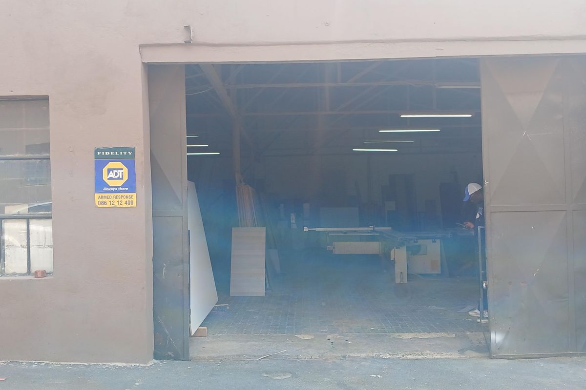 Industrial warehouse or workshop space featuring a large sliding metal entrance door, exposed roof trusses, and fluorescent lighting.
