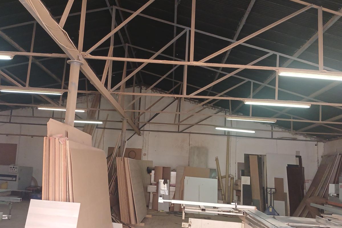 Industrial workshop space featuring exposed steel roof trusses, corrugated metal roofing, and overhead fluorescent lighting, currently utilized for material storage and manufacturing.
