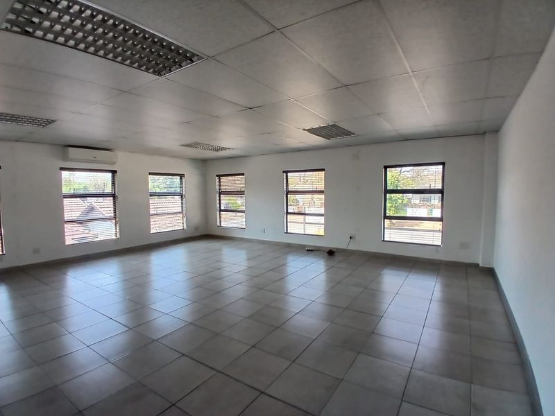A spacious, vacant office area featuring tiled flooring, a suspended ceiling with recessed lighting, and multiple windows providing natural light.