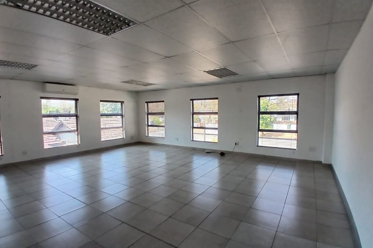 A spacious, vacant office area featuring tiled flooring, a suspended ceiling with recessed lighting, and multiple windows providing natural light.