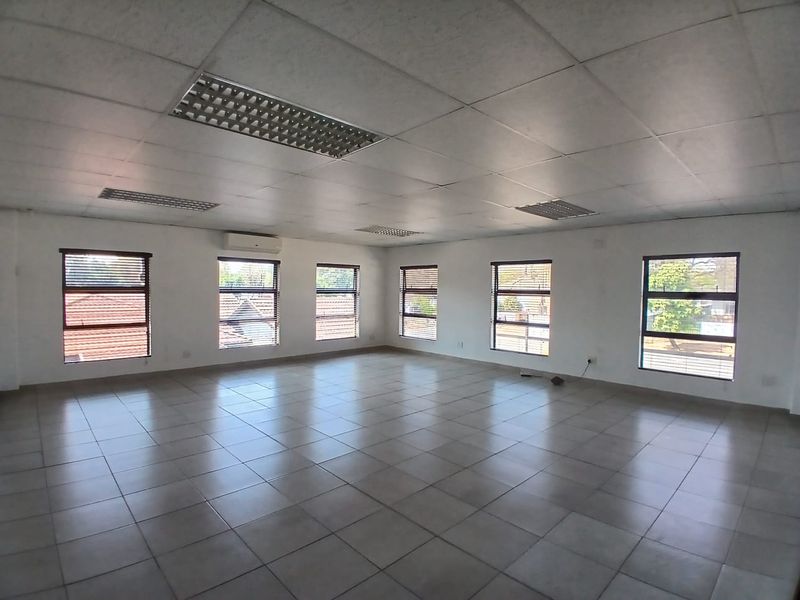A spacious, open-plan office area featuring tiled flooring, a suspended ceiling with recessed lighting, and multiple windows providing ample natural light.