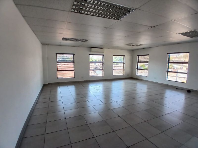 A spacious, empty office area featuring ceramic tiled flooring, a suspended ceiling with recessed lighting, and multiple windows equipped with venetian blinds.