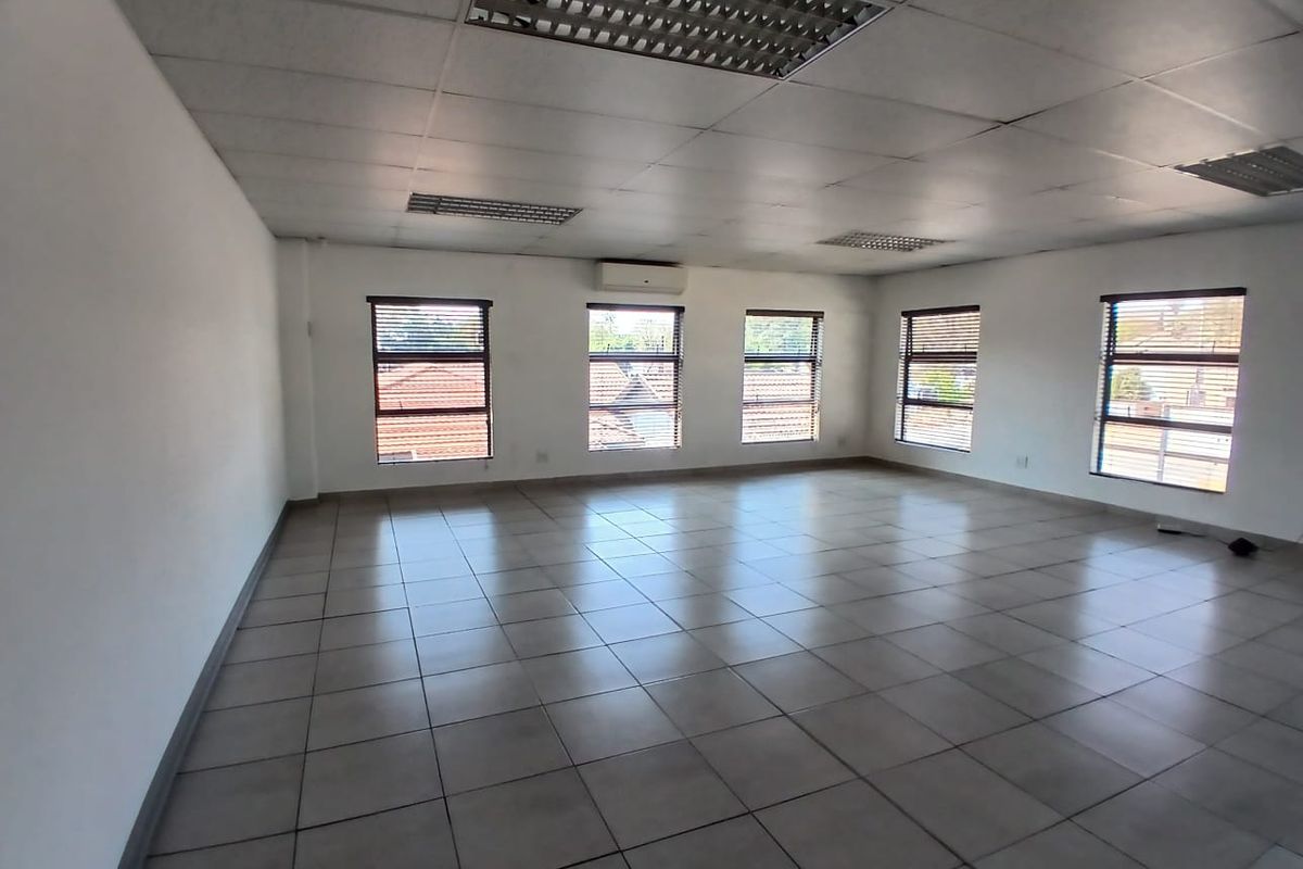 A spacious, empty office area featuring ceramic tiled flooring, a suspended ceiling with recessed lighting, and multiple windows equipped with venetian blinds.