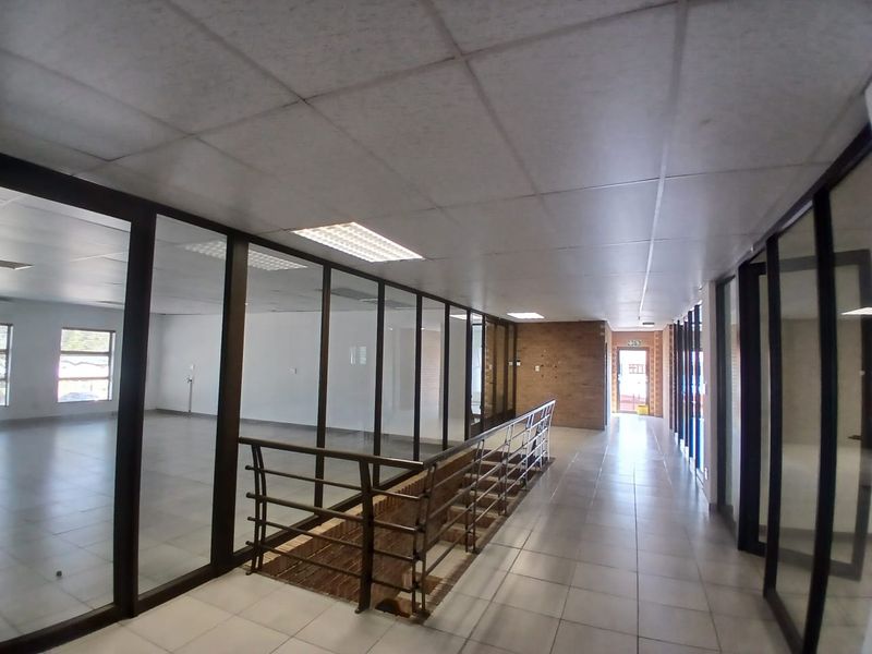Spacious office hallway featuring glass partitions, tiled flooring, suspended ceilings with recessed lighting, and a metal staircase railing overlooking the lower level.