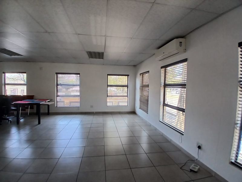 A spacious, open-plan office area featuring tiled flooring, a suspended ceiling, and multiple windows equipped with venetian blinds and air conditioning.