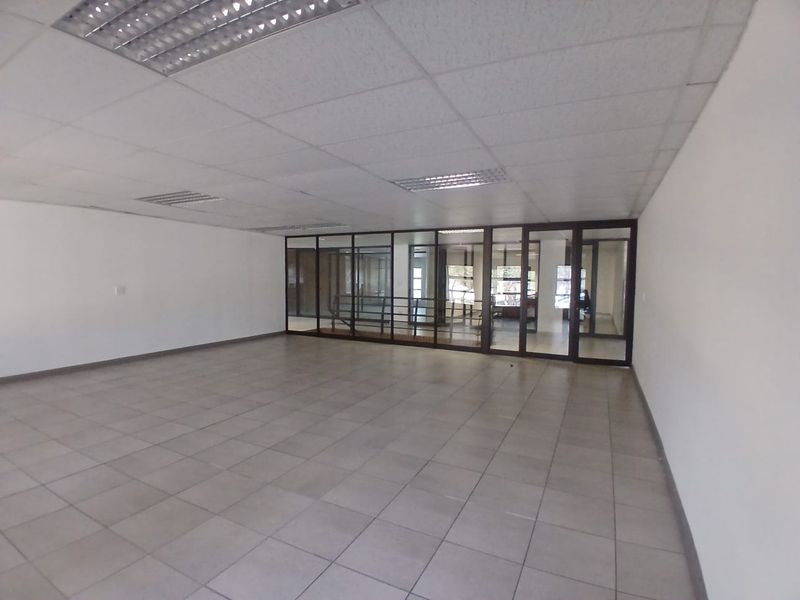 A spacious, empty office area featuring tiled flooring, a suspended ceiling with recessed lighting, and glass partition walls providing a modern, open-plan feel.
