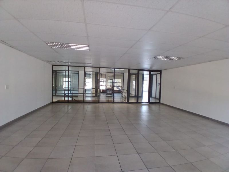 A spacious, empty open-plan office area featuring tiled flooring, a suspended ceiling with recessed lighting, and glass partition walls.