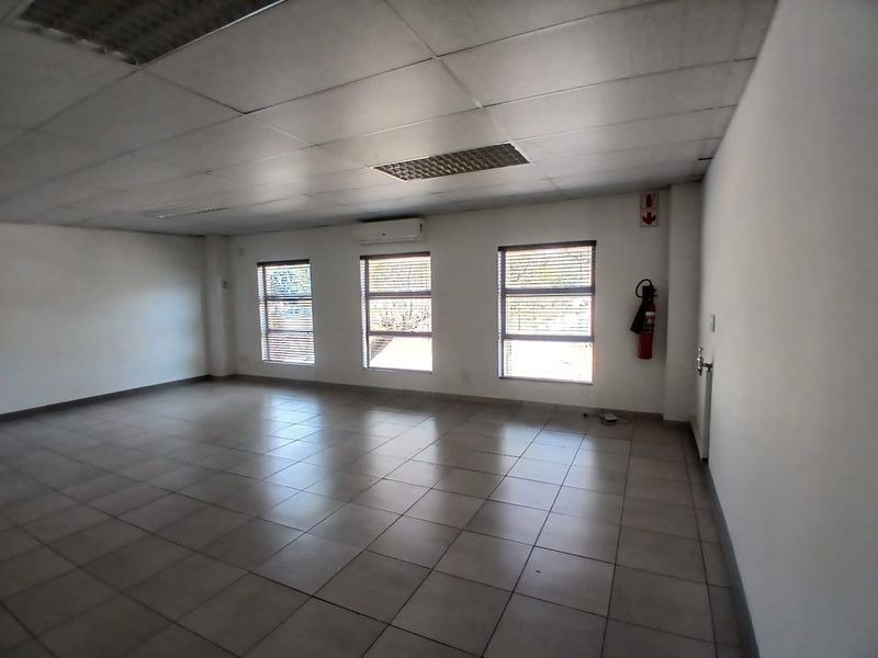 A spacious, empty office area featuring tiled flooring, a suspended ceiling with recessed lighting, and a wall-mounted air conditioning unit.