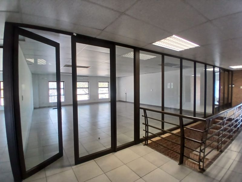 Spacious open-plan office area featuring glass partitions, tiled flooring, and a suspended ceiling with recessed lighting, overlooking a staircase.