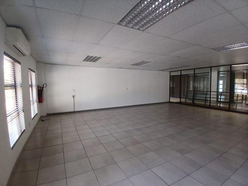 A spacious, empty office area featuring tiled flooring, a suspended ceiling with recessed lighting, and a glass partition wall.