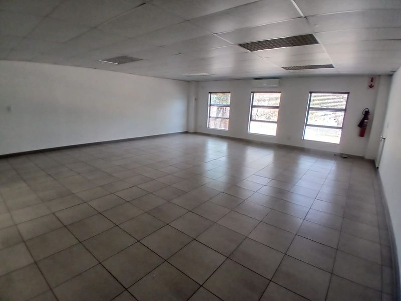 A spacious, empty office area featuring tiled flooring, a suspended ceiling with recessed lighting, and a wall-mounted air conditioning unit.