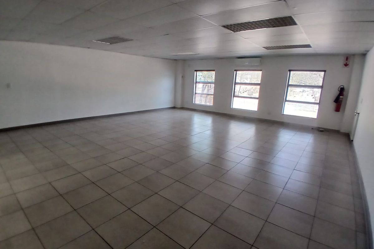 A spacious, empty office area featuring tiled flooring, a suspended ceiling with recessed lighting, and a wall-mounted air conditioning unit.