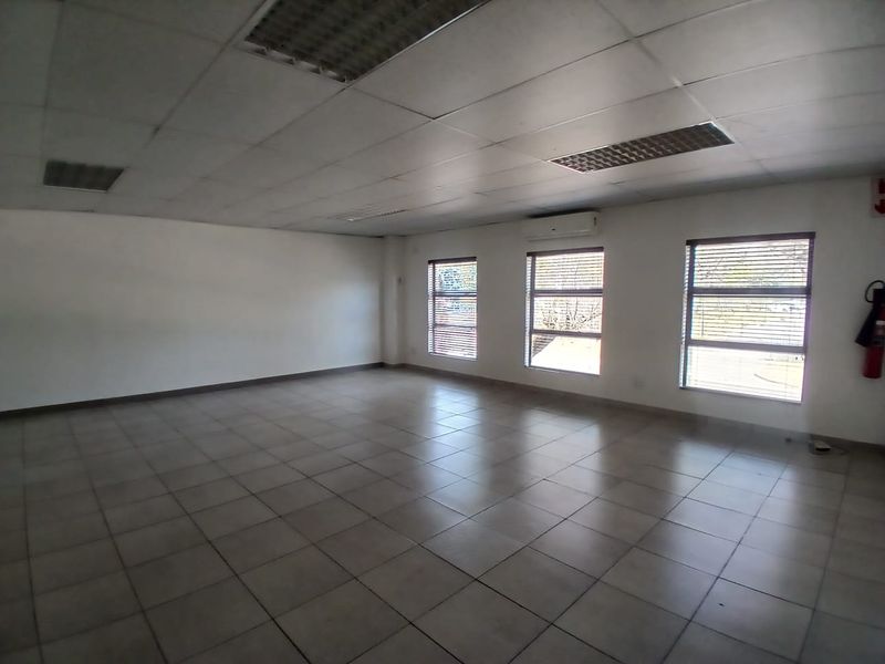 A spacious, empty office area featuring tiled flooring, a suspended ceiling with recessed lighting, and a wall-mounted air conditioning unit.
