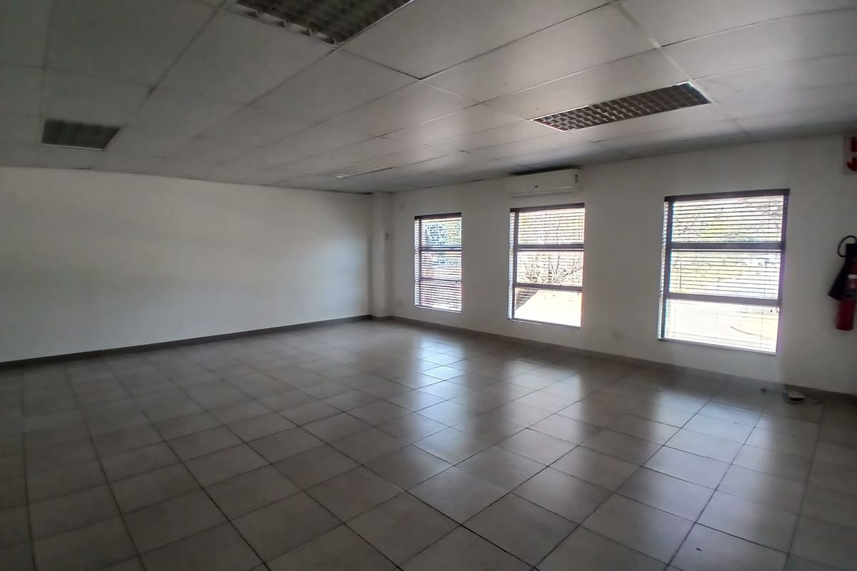 A spacious, empty office area featuring tiled flooring, a suspended ceiling with recessed lighting, and a wall-mounted air conditioning unit.