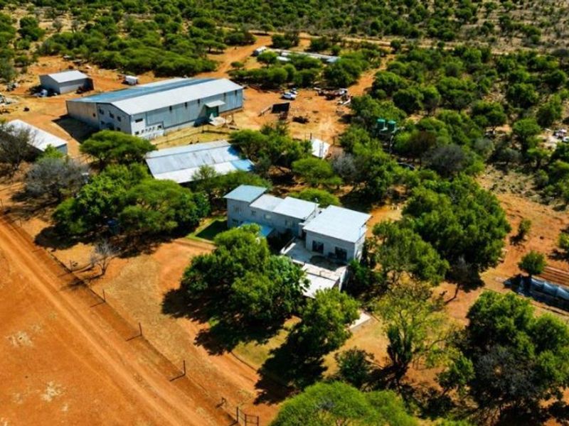Aerial view of a farm property featuring a large metal shed, main farmhouse, water storage tanks, and surrounding natural bushveld landscape.