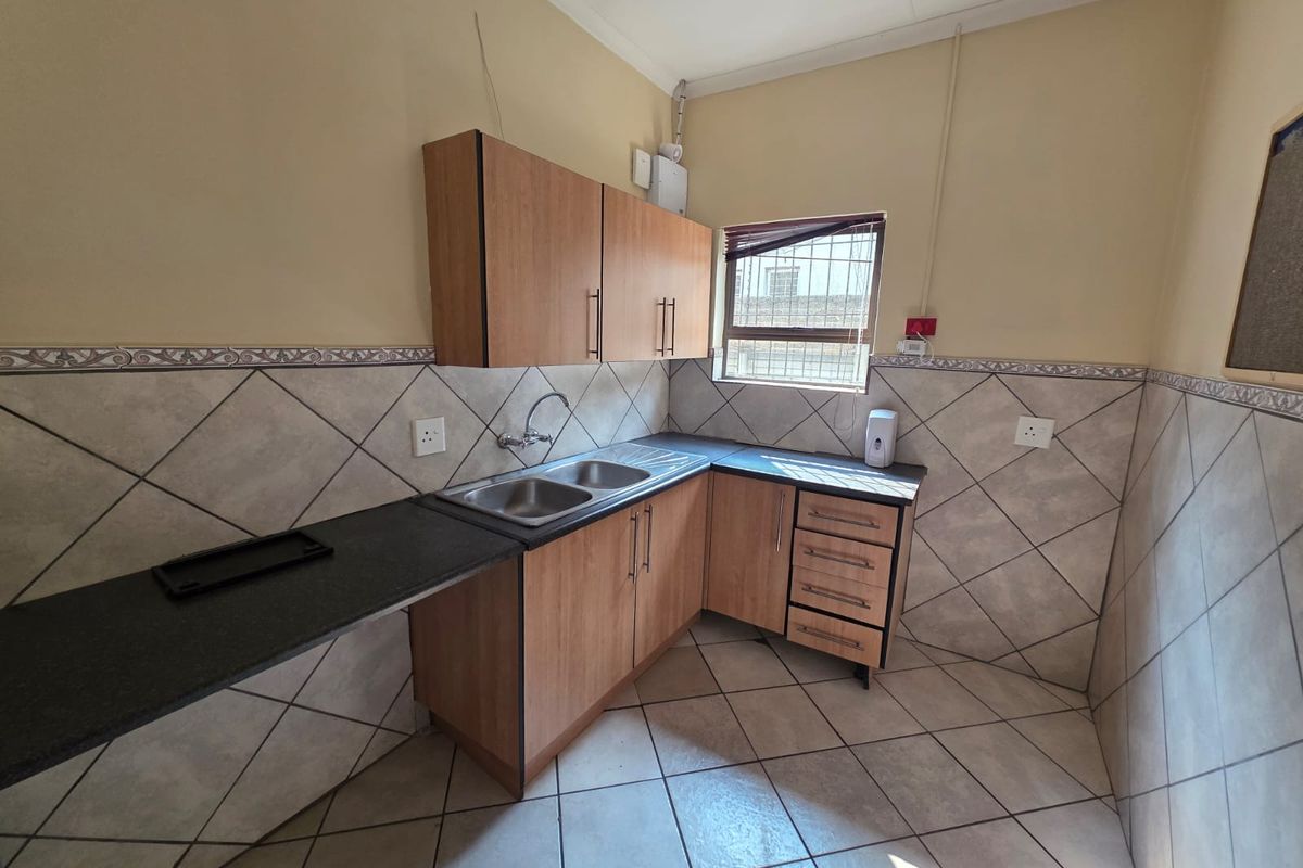 A functional kitchenette featuring wood-finish cabinetry, a double-basin sink, dark countertops, and tiled flooring with matching wall splashbacks.