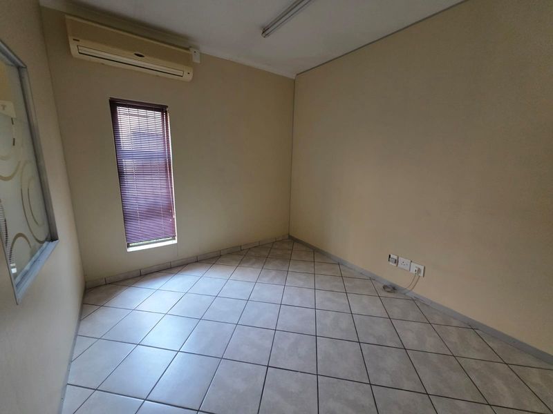 A vacant office space featuring tiled flooring, a wall-mounted air conditioning unit, and a window with fitted blinds.