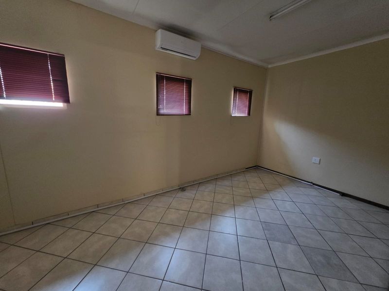 A functional office space featuring tiled flooring, wall-mounted air conditioning unit, and multiple windows with fitted blinds.