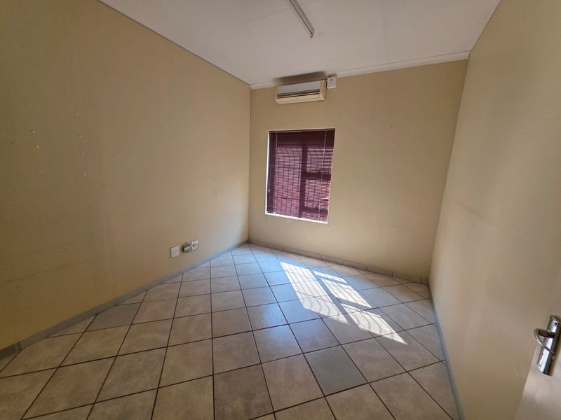 A vacant office space featuring tiled flooring, a wall-mounted air conditioning unit, and a window with fitted blinds.