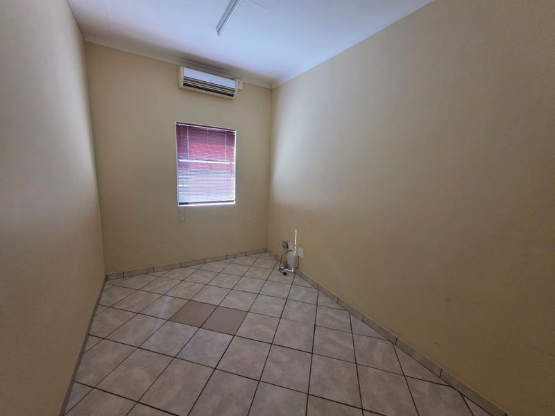 A compact, empty office space featuring tiled flooring, a wall-mounted air conditioning unit, and a window with fitted blinds.