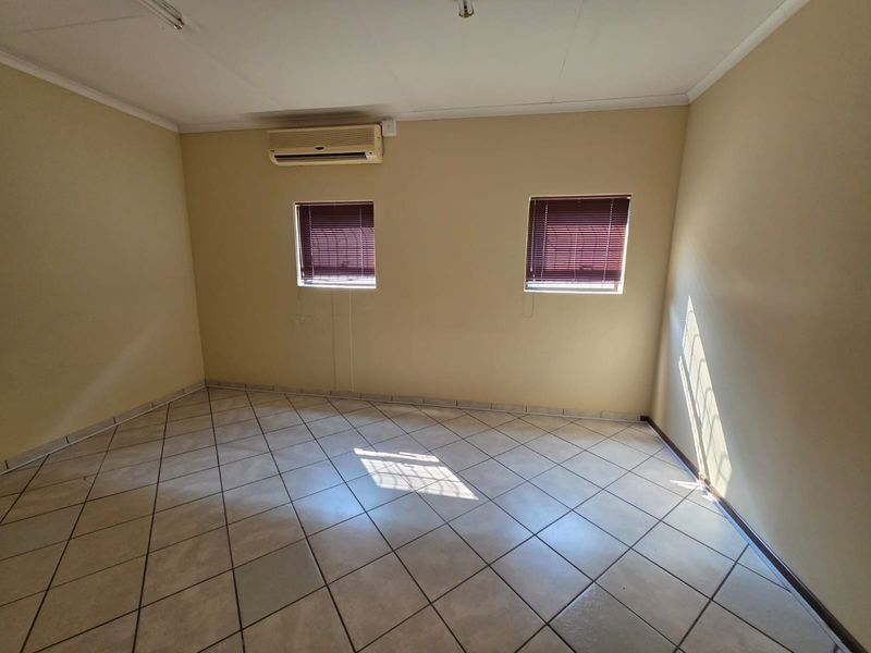 An empty office space featuring tiled flooring, two windows with fitted blinds, and a wall-mounted air conditioning unit.