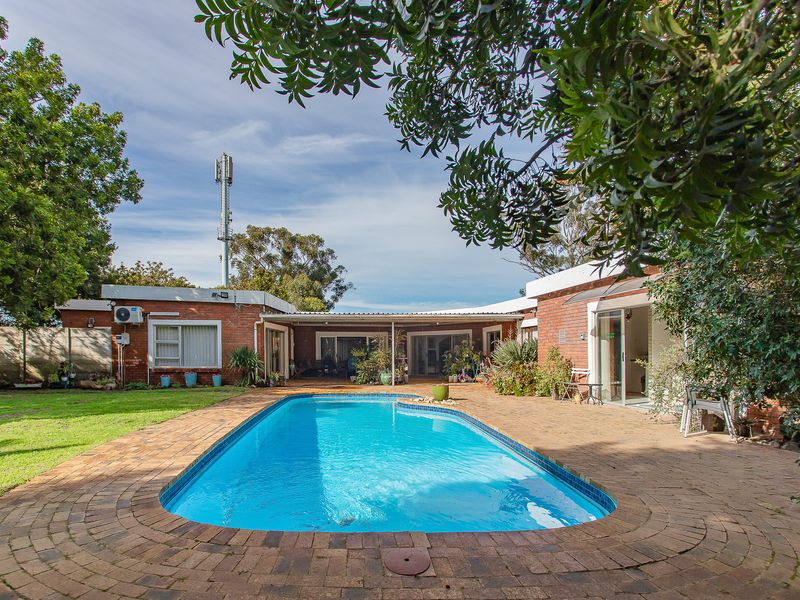 A spacious outdoor pool area featuring a swimming pool surrounded by brick paving, set against a brick farmhouse structure with multiple sliding door access points.