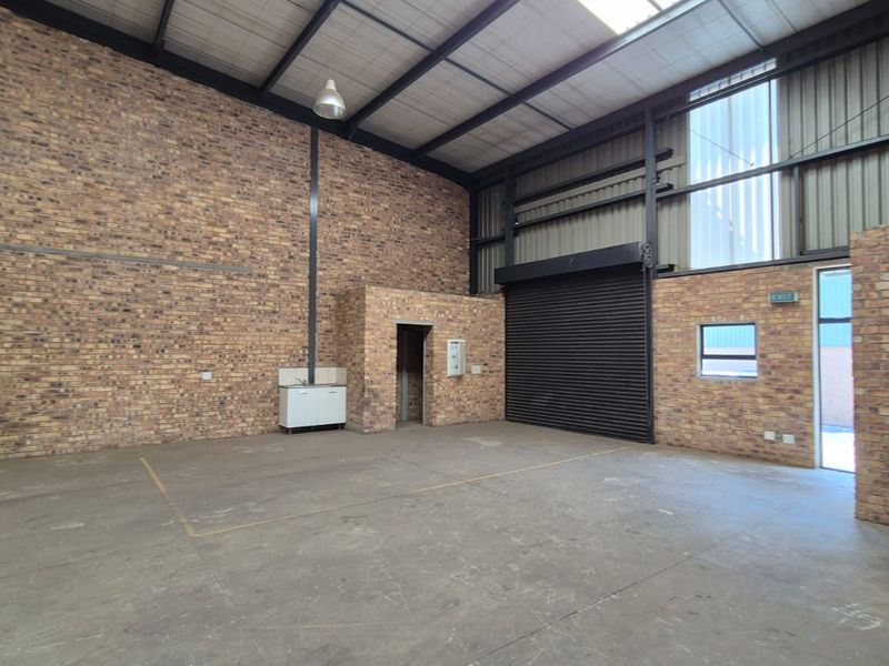Spacious industrial warehouse unit featuring exposed brick walls, concrete flooring, high ceilings with steel trusses, and a secure roller shutter door.