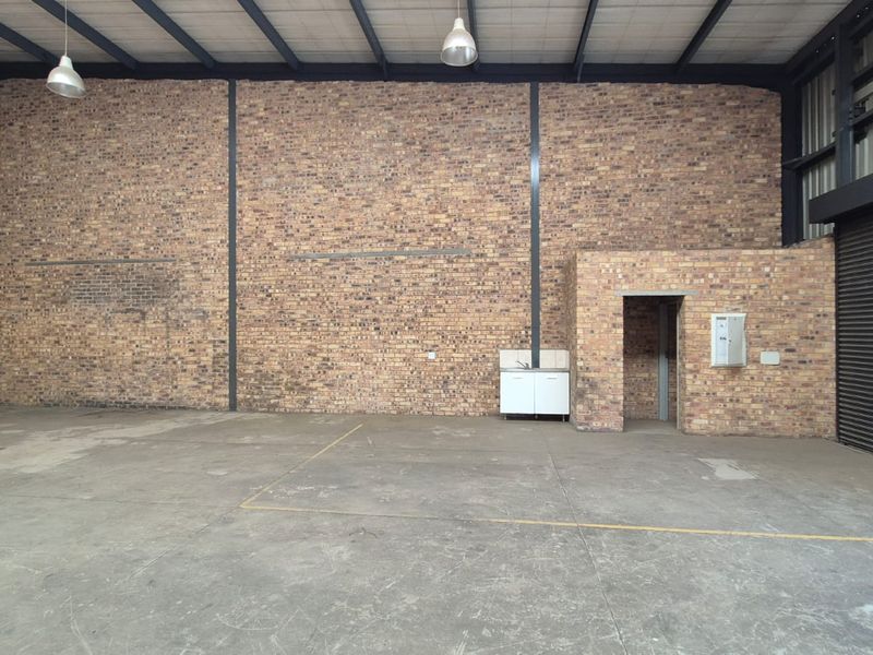 Spacious industrial warehouse unit featuring exposed brick walls, high steel truss ceilings, concrete flooring, a roller shutter door, and a small kitchenette area.