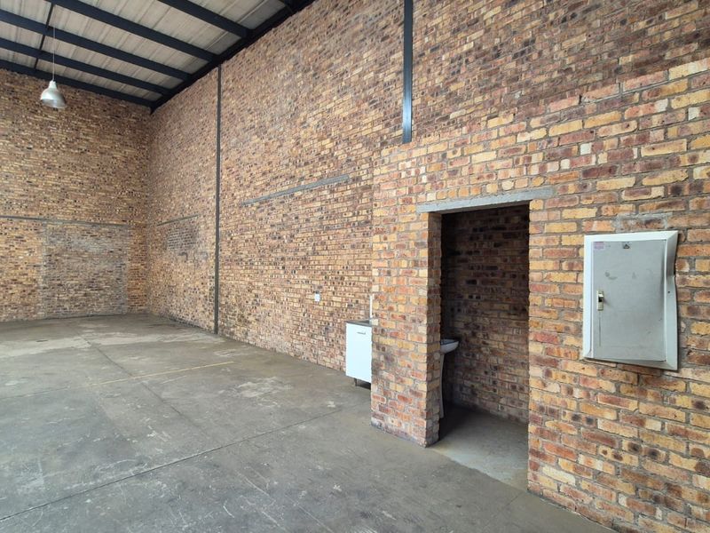 Spacious industrial utility area featuring exposed brick walls, concrete flooring, high steel roof trusses, and a small integrated washroom area.