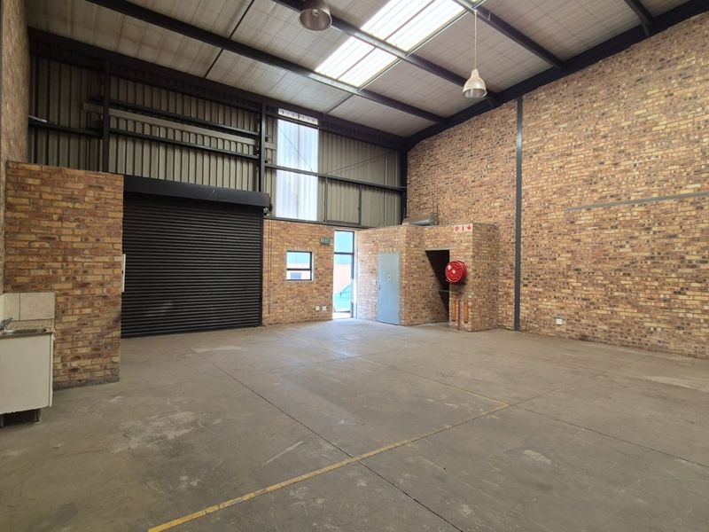 Spacious industrial warehouse unit featuring exposed brick walls, concrete flooring, a roller shutter door, and a small kitchenette area.