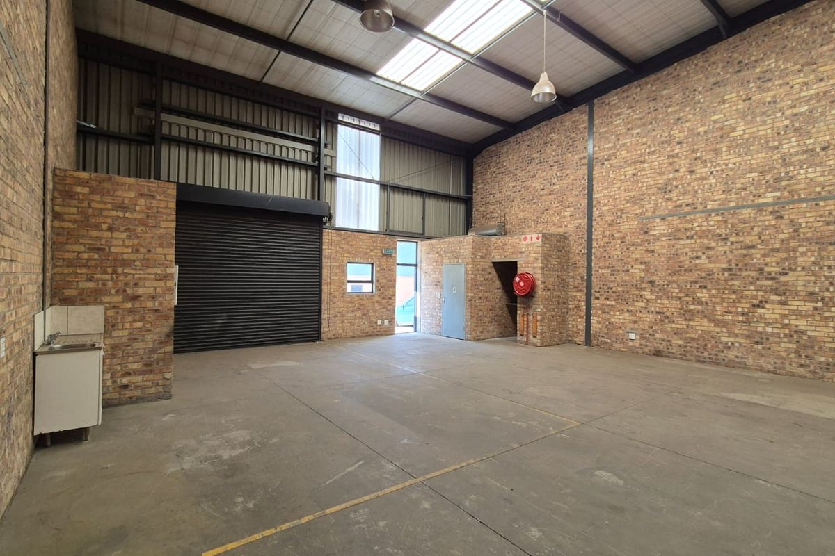 Spacious industrial warehouse unit featuring exposed brick walls, concrete flooring, a roller shutter door, and a small kitchenette area.