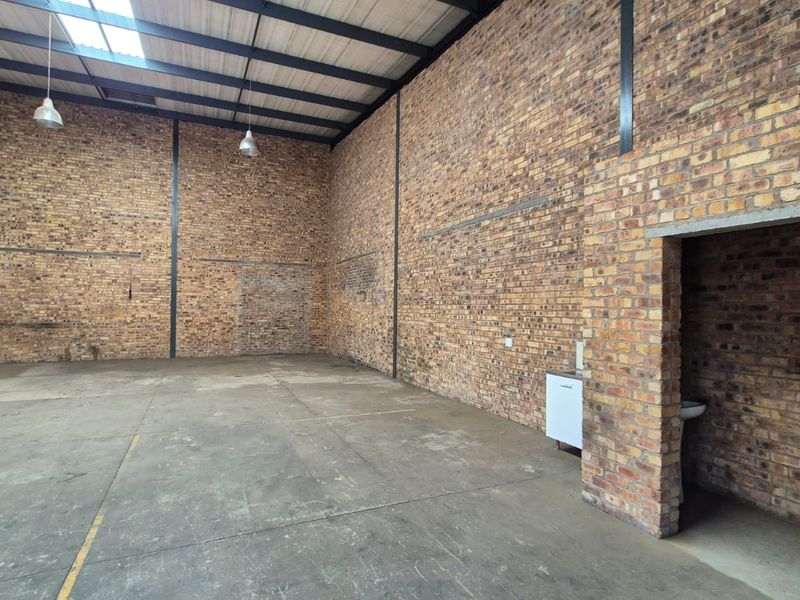 Spacious industrial warehouse unit featuring exposed brick walls, high ceilings with steel trusses, concrete flooring, and a small integrated washroom area.