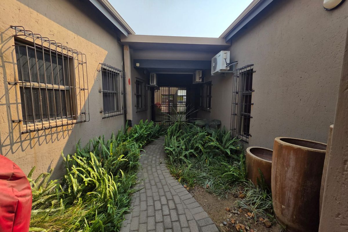 A paved walkway leads between two building wings, featuring landscaped garden beds, security bars on windows, and wall-mounted air conditioning units.