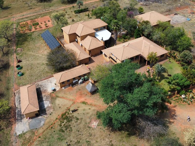 Aerial view of a farm property featuring multiple buildings with tiled roofs, a solar panel array, paved courtyard areas, and established garden surroundings.