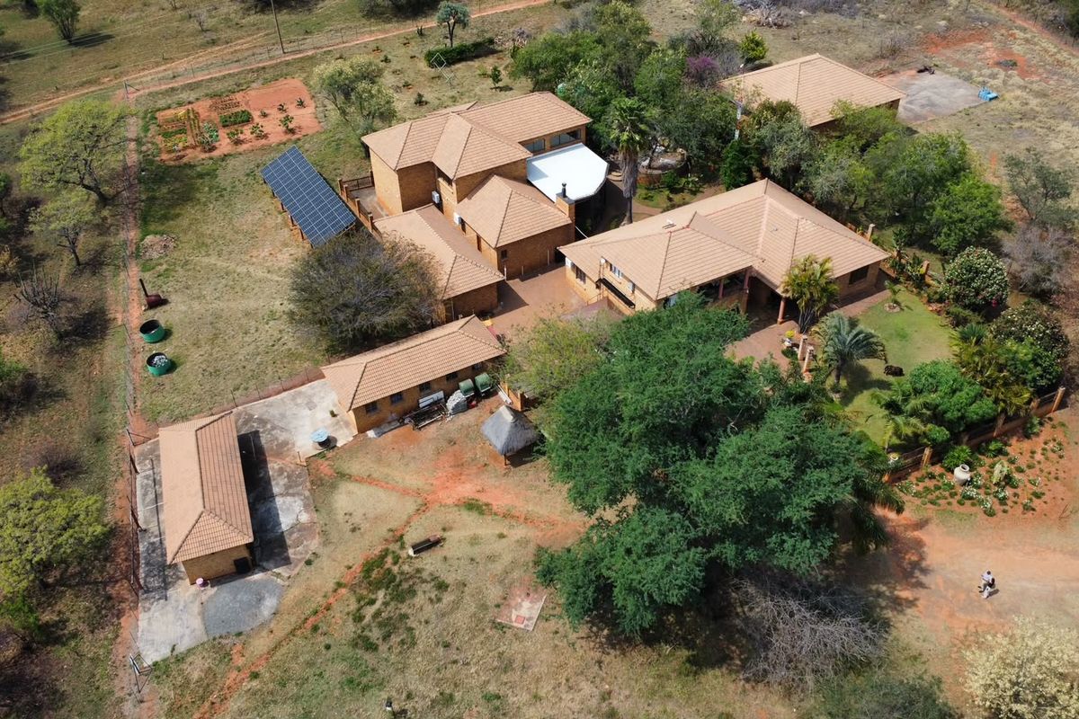 Aerial view of a farm property featuring multiple buildings with tiled roofs, a solar panel array, paved courtyard areas, and established garden surroundings.