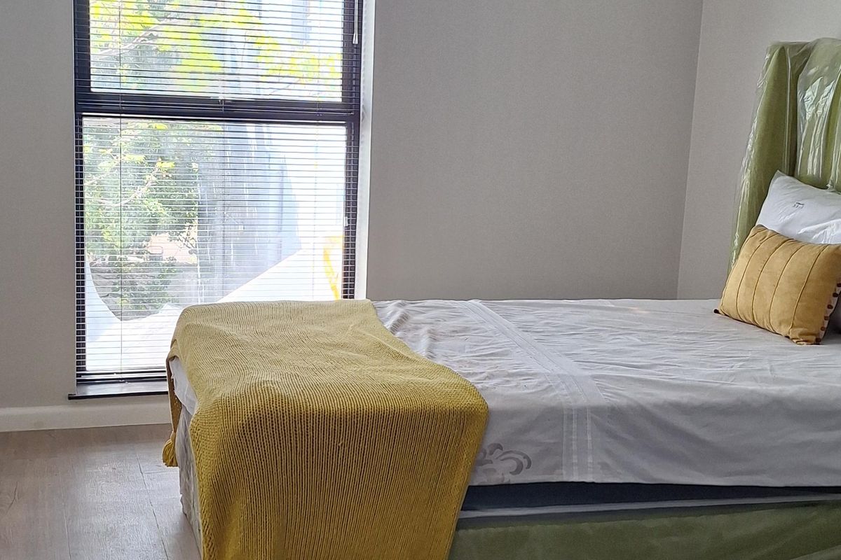 A bright and modern bedroom featuring light-toned laminate flooring, recessed ceiling lighting, and a large vertical window fitted with venetian blinds.
