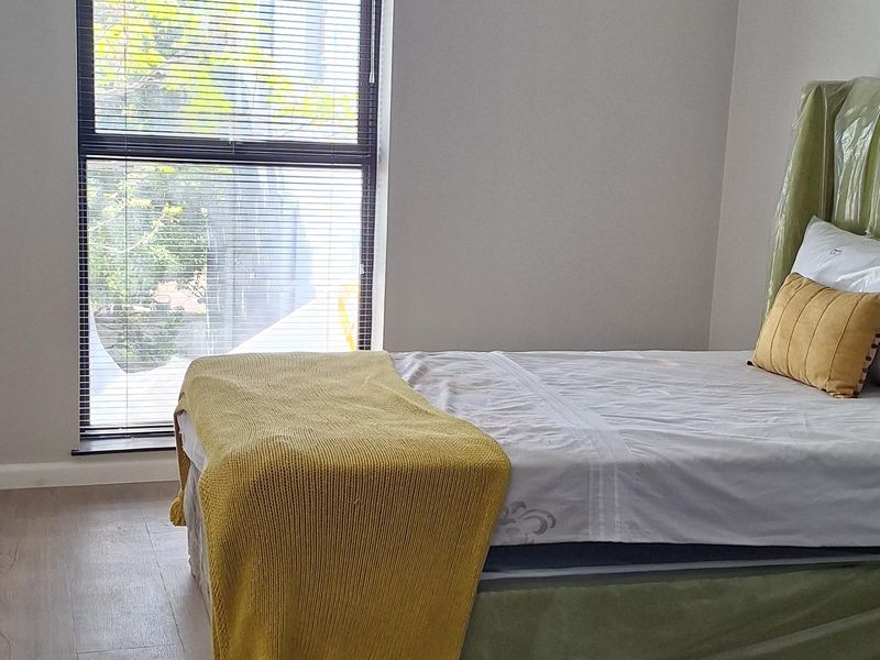 A bright and modern bedroom featuring light-toned laminate flooring, recessed ceiling lighting, and a large vertical window fitted with venetian blinds.