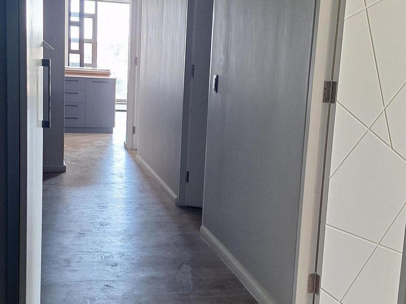 A bright, modern hallway featuring light-toned laminate flooring, neutral grey walls, and recessed downlighting, leading towards an open-plan area.
