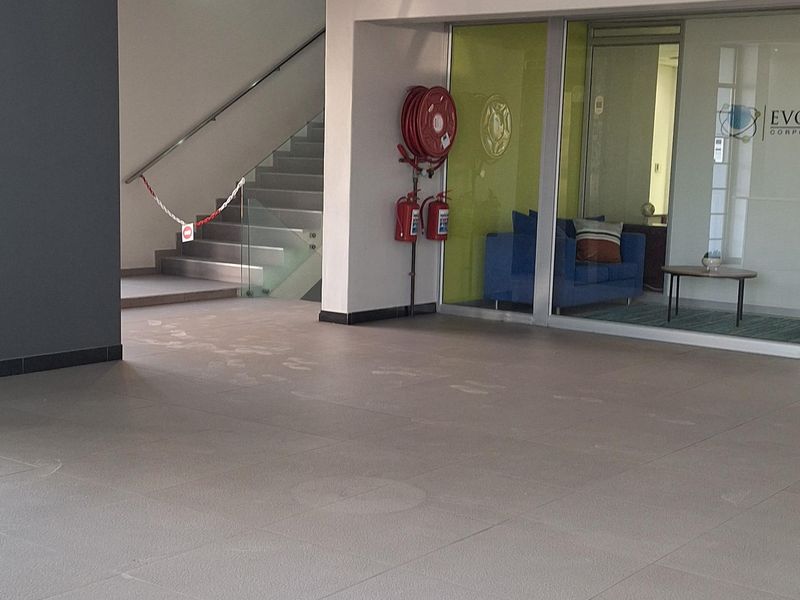 A spacious commercial building entrance featuring tiled flooring, recessed lighting, a staircase, and a glass-partitioned office area with integrated fire safety equipment.
