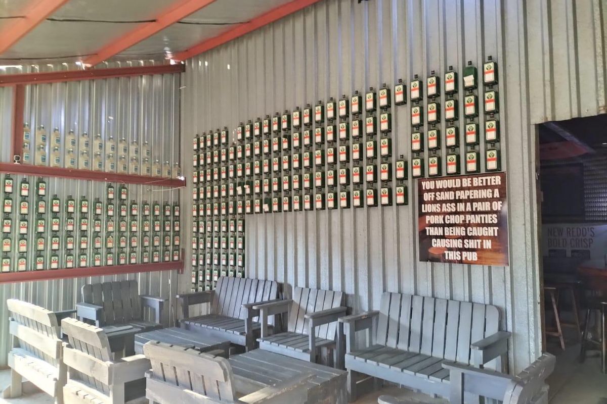 A rustic bar area featuring corrugated metal wall cladding and exposed steel roof beams, decorated with a wall-mounted collection of glass bottles.