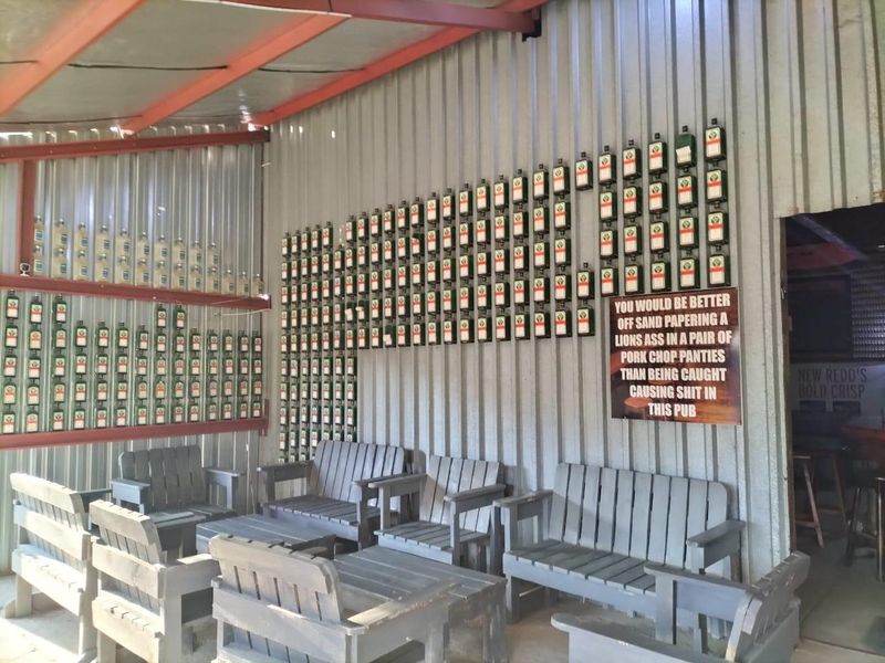 A rustic bar area featuring corrugated metal wall cladding and exposed steel roof beams, decorated with a wall-mounted collection of glass bottles.