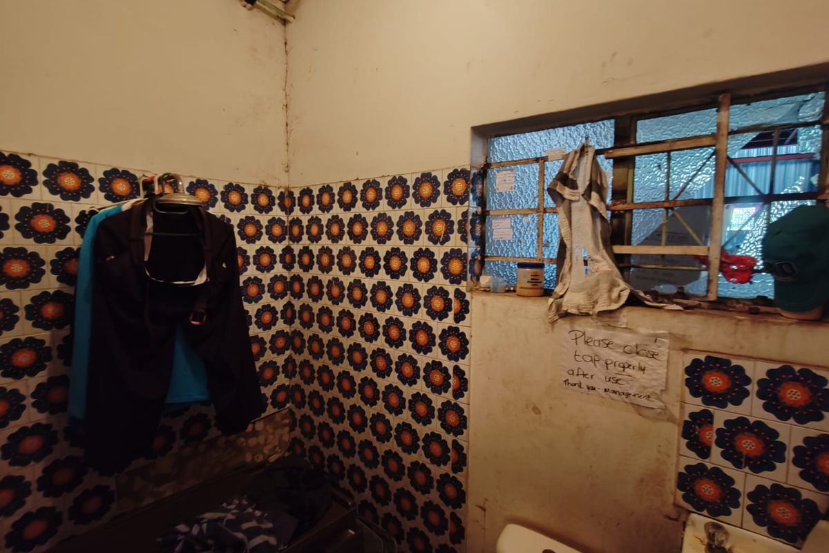 A dated bathroom featuring retro floral wall tiles, a standard toilet, and a window with a metal security frame.
