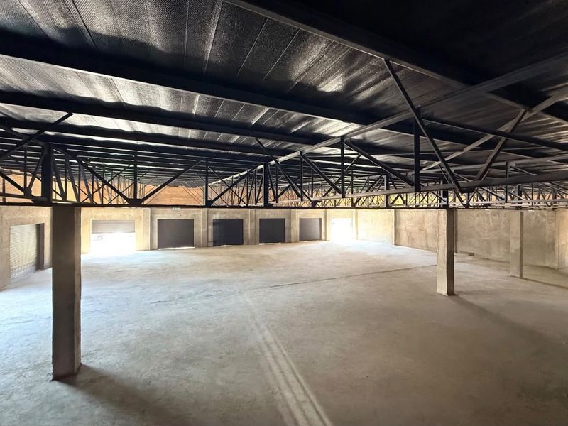 Spacious open-plan industrial utility area featuring exposed steel roof trusses, concrete flooring, and multiple roller shutter doors, suitable for storage or workshop use.